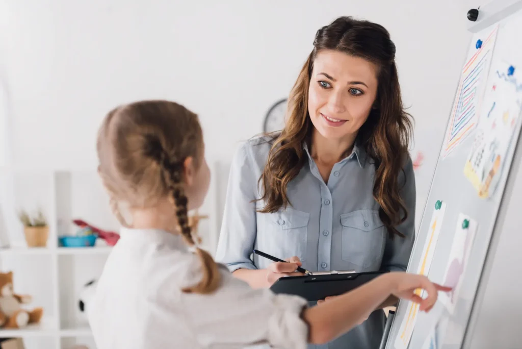 Teacher explains colorful charts on a whiteboard to a student in a bright classroom, holding a clipboard and pen.