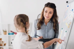 Teacher explains colorful charts on a whiteboard to a student in a bright classroom, holding a clipboard and pen.